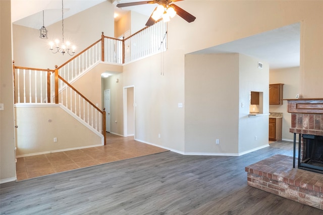 unfurnished living room featuring a towering ceiling, a brick fireplace, ceiling fan with notable chandelier, and hardwood / wood-style floors