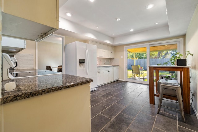 kitchen with white appliances, backsplash, dark stone countertops, a tray ceiling, and white cabinetry