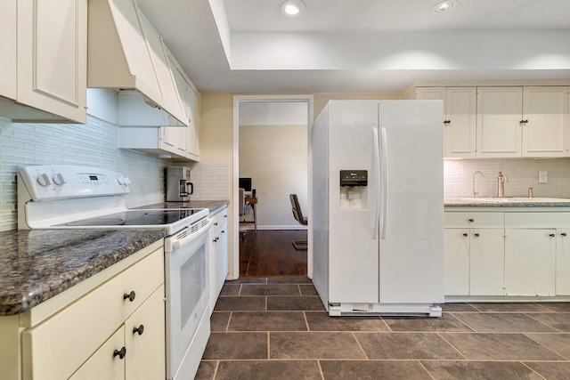 kitchen with custom range hood, white appliances, sink, dark stone countertops, and cream cabinetry