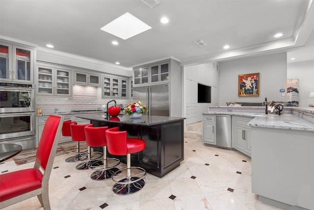 kitchen featuring a kitchen island with sink, gray cabinetry, stainless steel appliances, light stone countertops, and tasteful backsplash