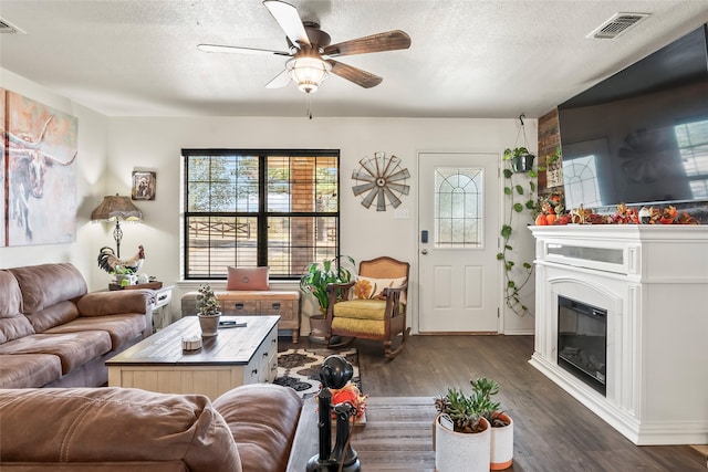 living room featuring a textured ceiling, ceiling fan, dark wood-style flooring, visible vents, and a glass covered fireplace