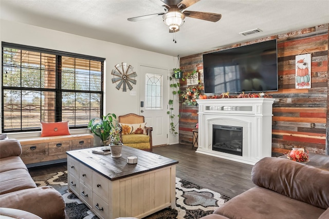 living room featuring wood walls, dark wood-style flooring, visible vents, a ceiling fan, and a glass covered fireplace