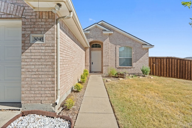 entrance to property with brick siding, a lawn, an attached garage, and fence