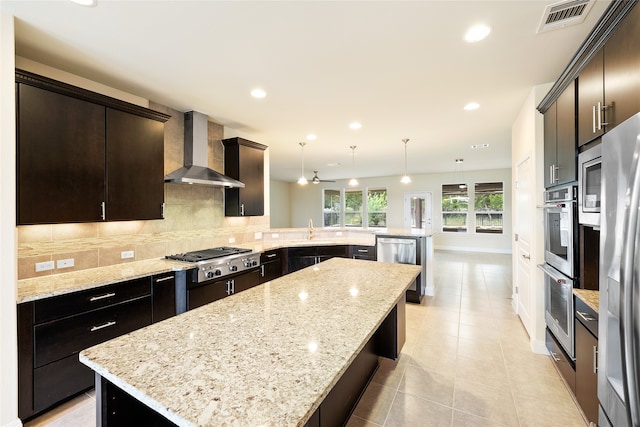 kitchen featuring light stone counters, wall chimney exhaust hood, stainless steel appliances, a kitchen island, and hanging light fixtures