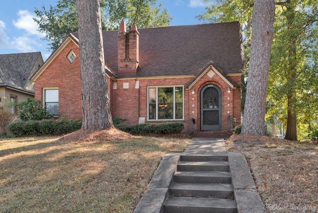 english style home featuring roof with shingles, a front yard, a chimney, and brick siding