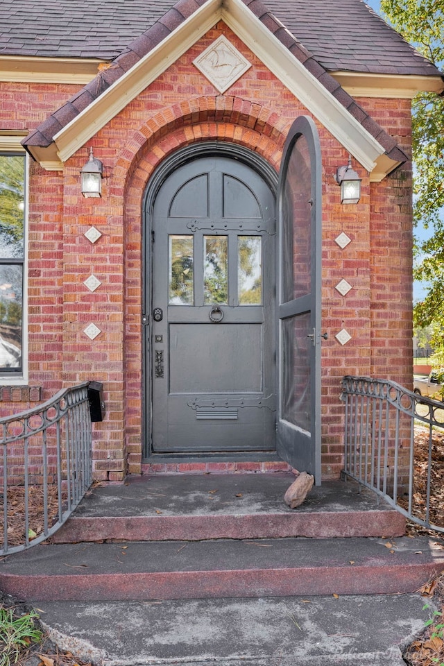 view of exterior entry with brick siding and roof with shingles