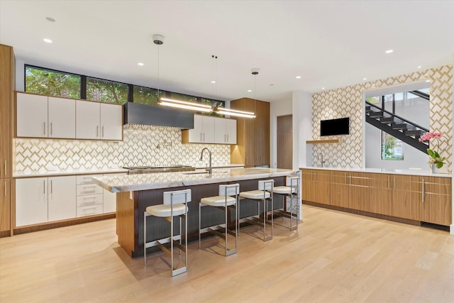 kitchen with pendant lighting, a center island with sink, wall chimney range hood, a kitchen bar, and white cabinetry