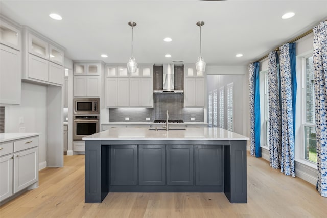 kitchen with wall chimney range hood, a kitchen island with sink, light hardwood / wood-style flooring, pendant lighting, and stainless steel appliances