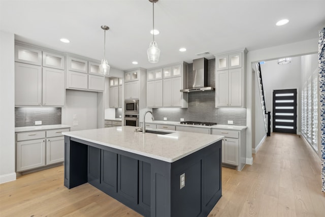 kitchen with wall chimney range hood, sink, appliances with stainless steel finishes, light hardwood / wood-style floors, and a center island with sink