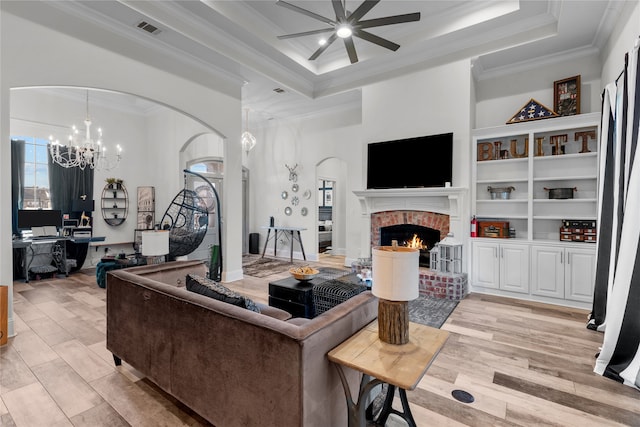 living room featuring ornamental molding, a brick fireplace, light wood-type flooring, a raised ceiling, and ceiling fan with notable chandelier