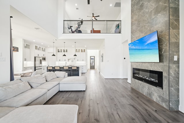 living room featuring a towering ceiling, wood-type flooring, a tiled fireplace, ceiling fan, and tile walls