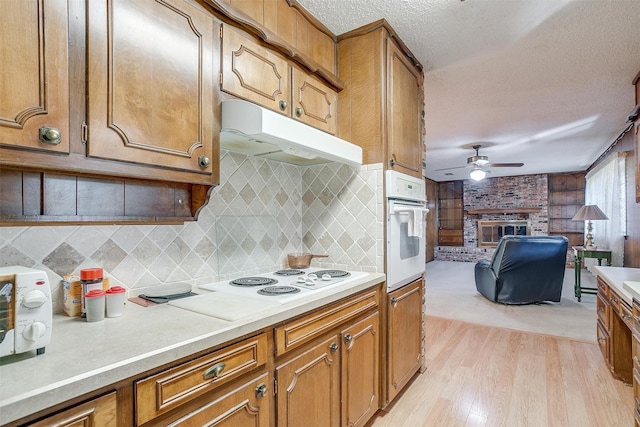 kitchen with backsplash, a brick fireplace, light wood-type flooring, white appliances, and ceiling fan