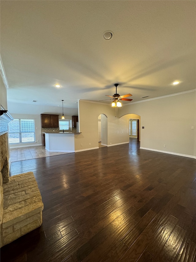 unfurnished living room featuring dark wood-type flooring, ceiling fan, a stone fireplace, and crown molding