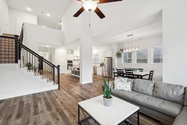 living room featuring stairway, a high ceiling, wood finished floors, baseboards, and ceiling fan with notable chandelier