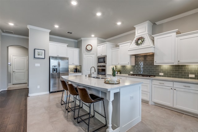 kitchen featuring tasteful backsplash, custom exhaust hood, an island with sink, stainless steel appliances, and white cabinets