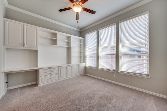 unfurnished office featuring built in desk, light colored carpet, ceiling fan, and crown molding