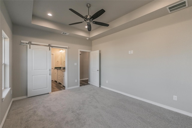 unfurnished bedroom featuring a tray ceiling, a barn door, ensuite bathroom, carpet floors, and ceiling fan