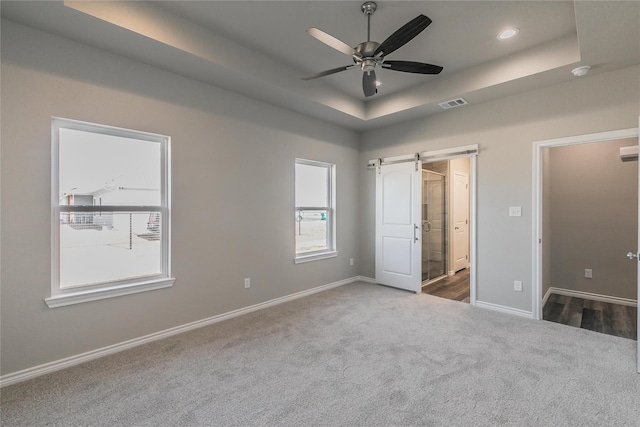 unfurnished bedroom with ceiling fan, a barn door, carpet flooring, a tray ceiling, and ensuite bathroom