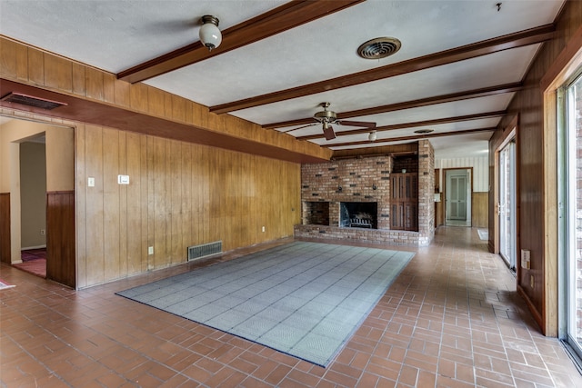 unfurnished living room featuring ceiling fan, a textured ceiling, a brick fireplace, beamed ceiling, and wooden walls