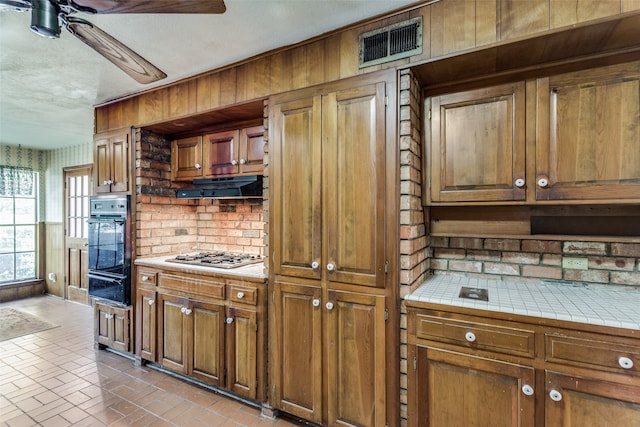 kitchen with ceiling fan, backsplash, stainless steel gas stovetop, black oven, and tile counters