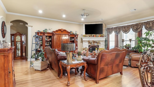 living room with a stone fireplace, light hardwood / wood-style flooring, ornamental molding, decorative columns, and ceiling fan