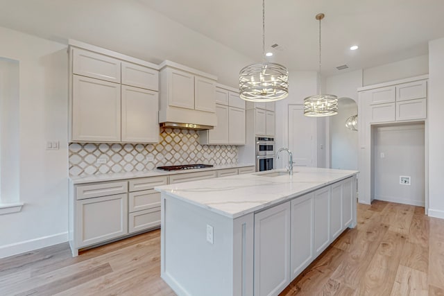 kitchen featuring stainless steel appliances, a center island with sink, pendant lighting, light hardwood / wood-style floors, and tasteful backsplash