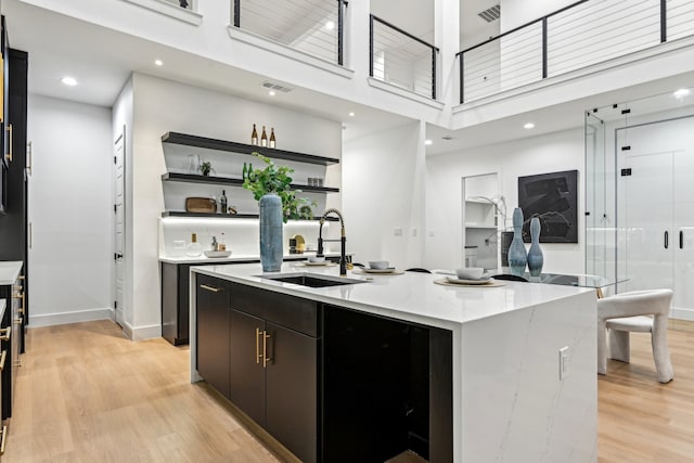 kitchen with a high ceiling, a kitchen island with sink, sink, light stone countertops, and light wood-type flooring