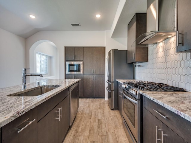 kitchen with light wood finished floors, a sink, dark brown cabinets, appliances with stainless steel finishes, and wall chimney exhaust hood