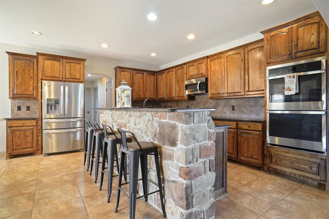 kitchen featuring light tile patterned floors, a breakfast bar area, backsplash, stainless steel appliances, and a center island with sink