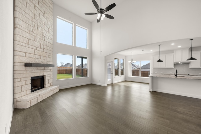 unfurnished living room featuring a towering ceiling, ceiling fan, dark wood-type flooring, sink, and a stone fireplace