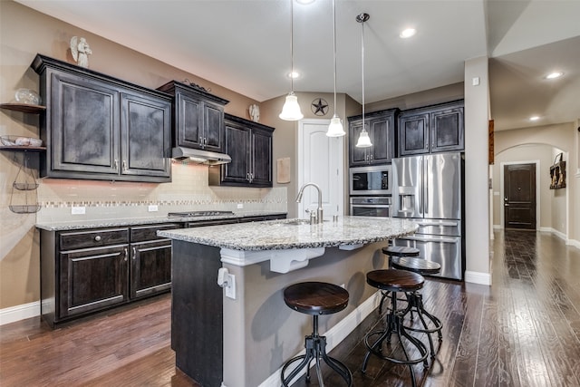 kitchen with a sink, under cabinet range hood, appliances with stainless steel finishes, arched walkways, and dark wood-style flooring