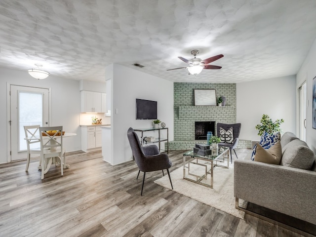 living room with ceiling fan, a textured ceiling, a brick fireplace, and light wood-type flooring