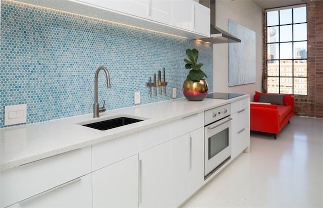 kitchen featuring black electric stovetop, wall chimney exhaust hood, white oven, sink, and white cabinetry