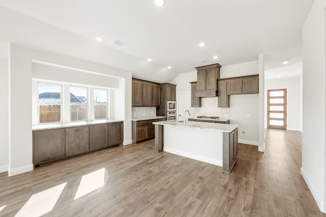 kitchen with light stone counters, vaulted ceiling, a center island with sink, light wood-type flooring, and decorative backsplash