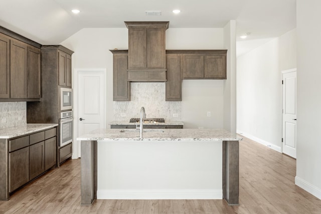 kitchen featuring appliances with stainless steel finishes, light stone countertops, an island with sink, and backsplash