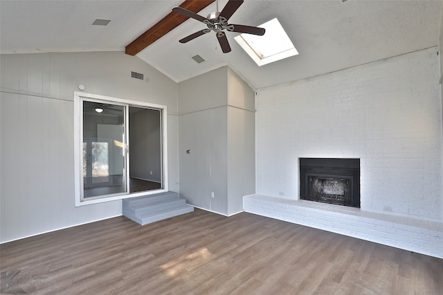 unfurnished living room with wood-type flooring, vaulted ceiling with beams, a fireplace, ceiling fan, and brick wall