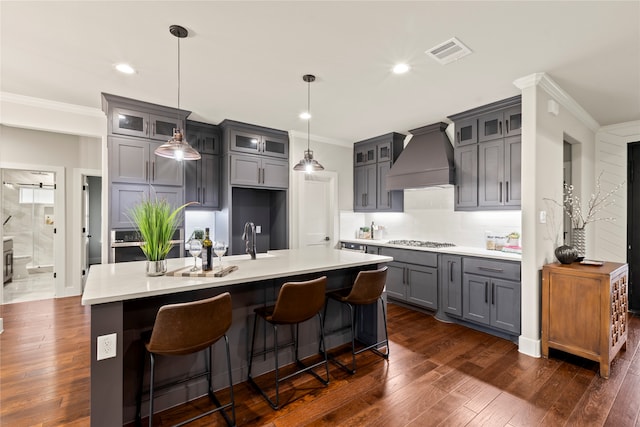 kitchen with custom range hood, a center island with sink, hanging light fixtures, and dark wood-type flooring