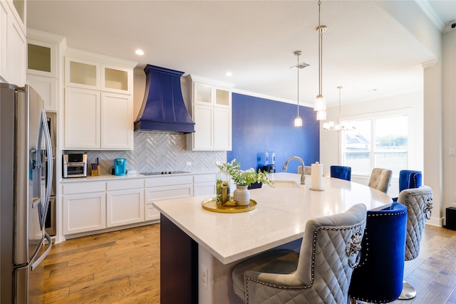 kitchen featuring stainless steel fridge, hanging light fixtures, a center island with sink, premium range hood, and light wood-type flooring