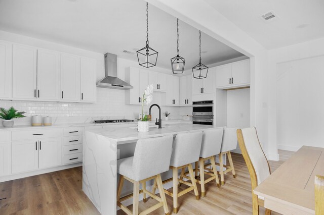 kitchen with backsplash, a kitchen island with sink, wall chimney range hood, pendant lighting, and white cabinetry