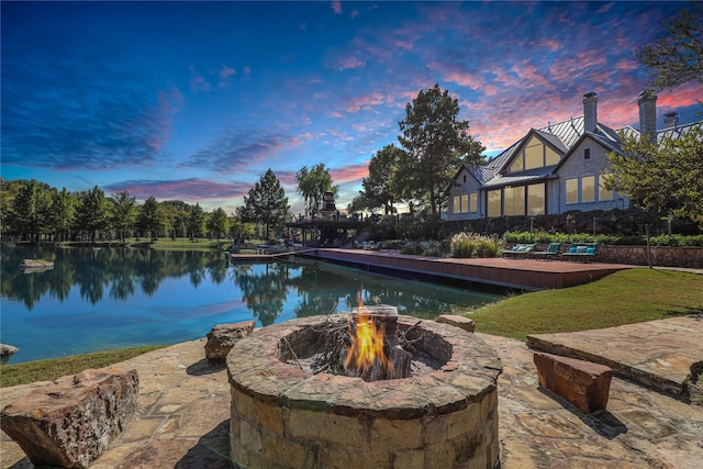 pool at dusk featuring a patio, a yard, a water view, and a fire pit