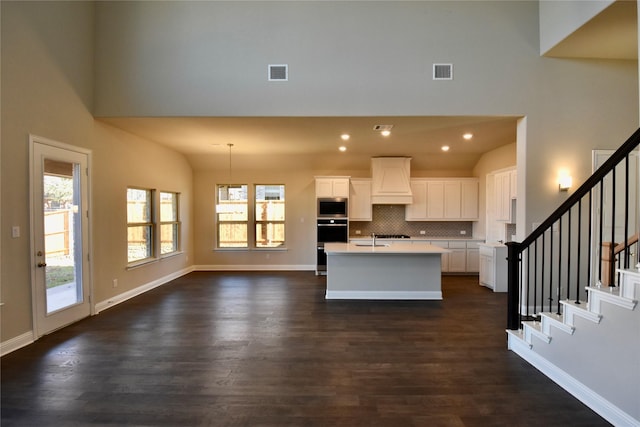 kitchen with stainless steel microwave, backsplash, a kitchen island with sink, premium range hood, and white cabinetry