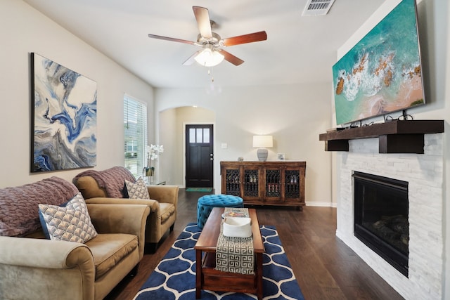 living room featuring dark wood-type flooring, ceiling fan, and a stone fireplace