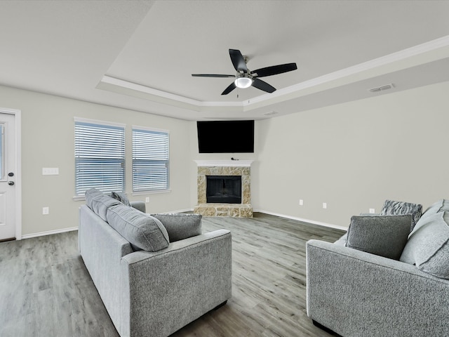 living room featuring light hardwood / wood-style floors, a stone fireplace, a raised ceiling, and ceiling fan