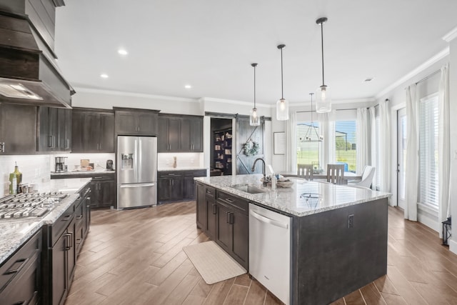 kitchen with sink, wall chimney range hood, hardwood / wood-style floors, an island with sink, and appliances with stainless steel finishes