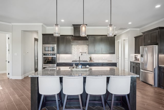 kitchen featuring appliances with stainless steel finishes, custom exhaust hood, dark hardwood / wood-style floors, hanging light fixtures, and an island with sink