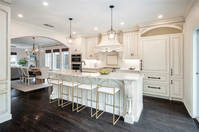 kitchen with light stone countertops, cream cabinetry, an island with sink, pendant lighting, and dark hardwood / wood-style floors