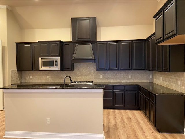 kitchen featuring tasteful backsplash, stainless steel microwave, light hardwood / wood-style floors, dark stone countertops, and premium range hood