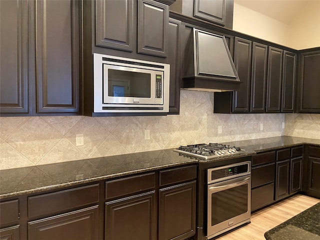 kitchen featuring appliances with stainless steel finishes, backsplash, dark stone counters, light hardwood / wood-style flooring, and premium range hood