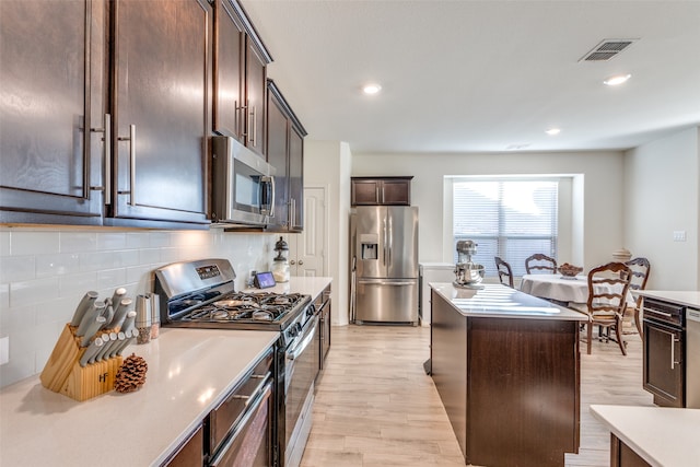 kitchen with a kitchen island, backsplash, dark brown cabinetry, light wood-type flooring, and appliances with stainless steel finishes