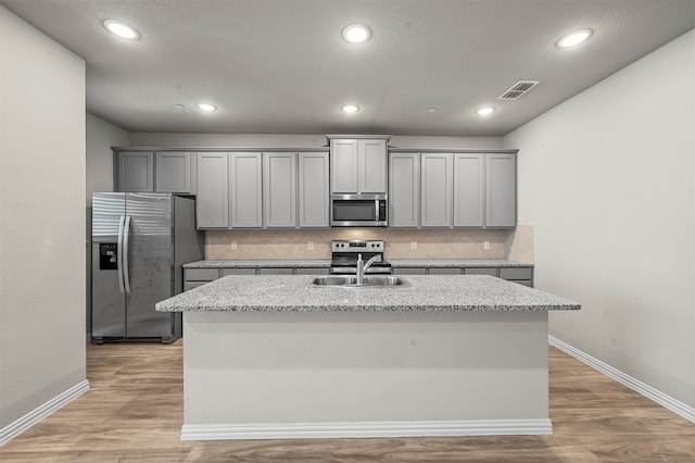 kitchen with stainless steel appliances, light stone countertops, a center island with sink, and gray cabinets